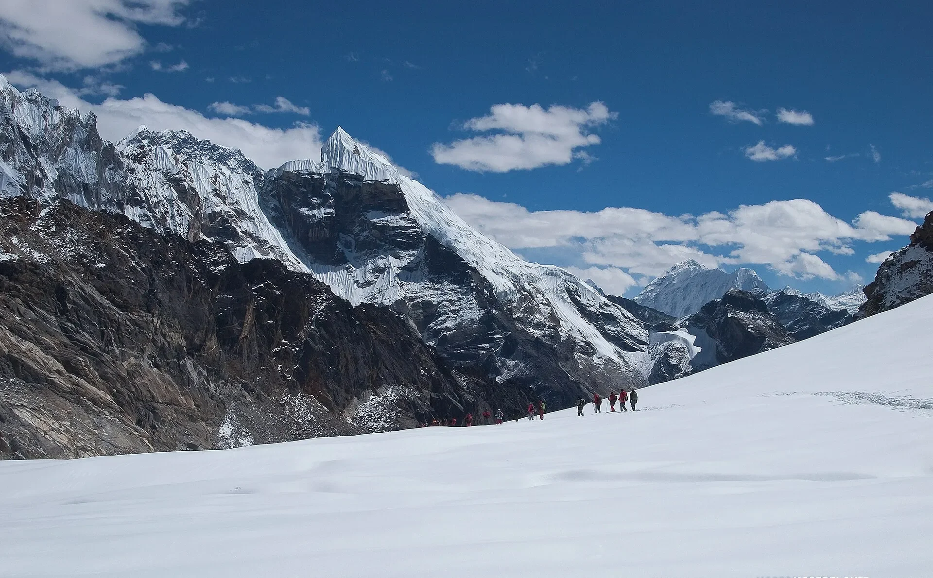 A line of tiny trekkers navigating across a vast, snow-covered glacier field toward dark, rugged mountain ridges under a blue sky
