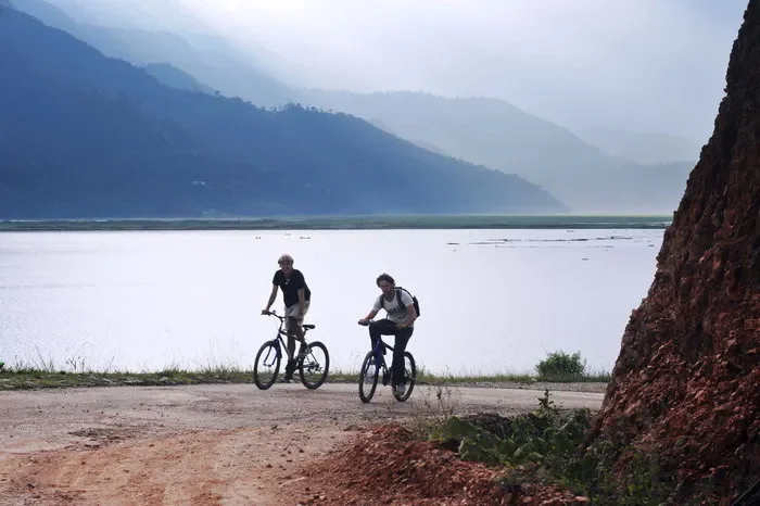 wo cyclists riding mountain bikes along a paved trail next to Phewa Lake with misty hills in the background.
