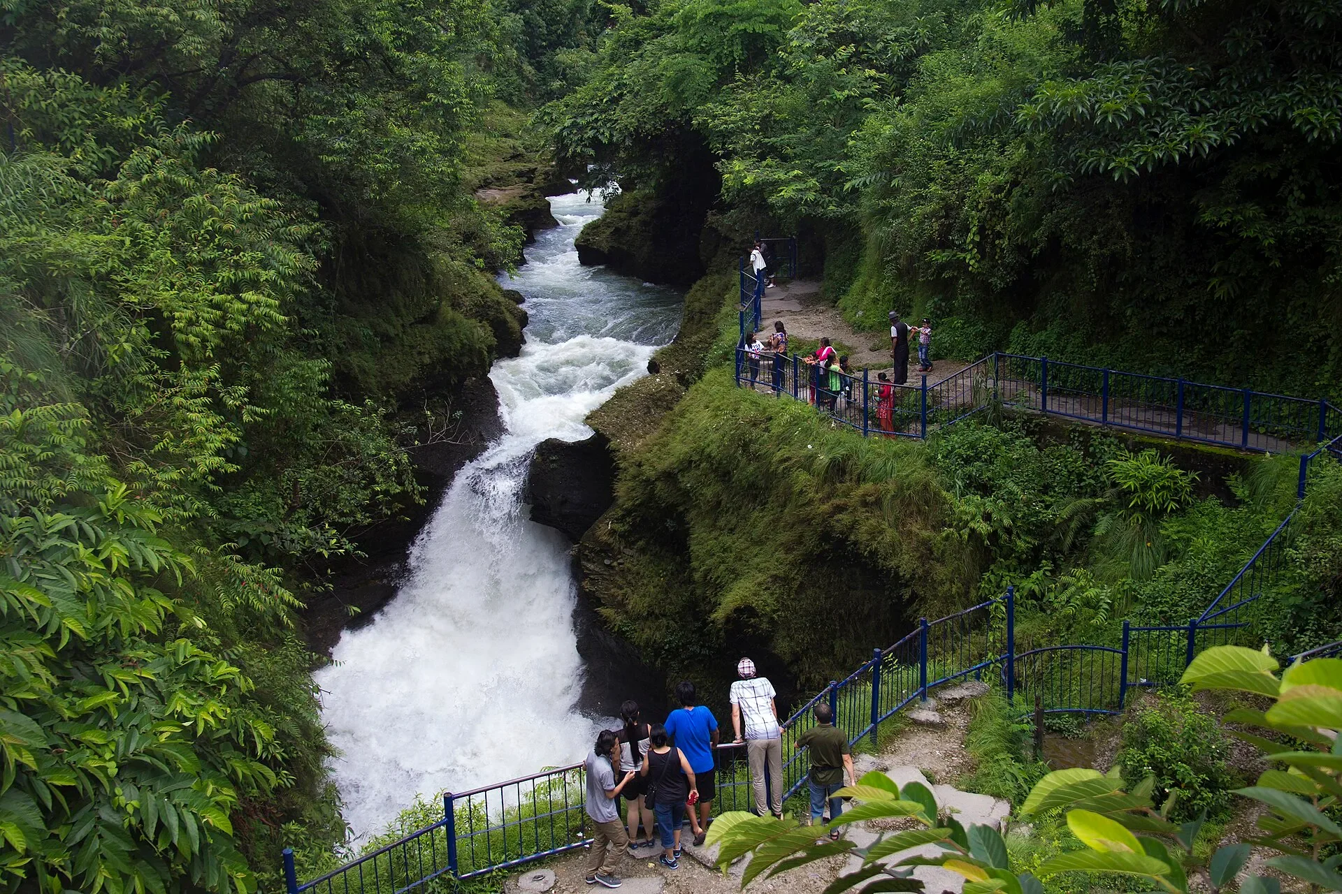 Visitors standing along a fenced walkway watching the rushing water plunge into Davis Falls (Patale Chhango).