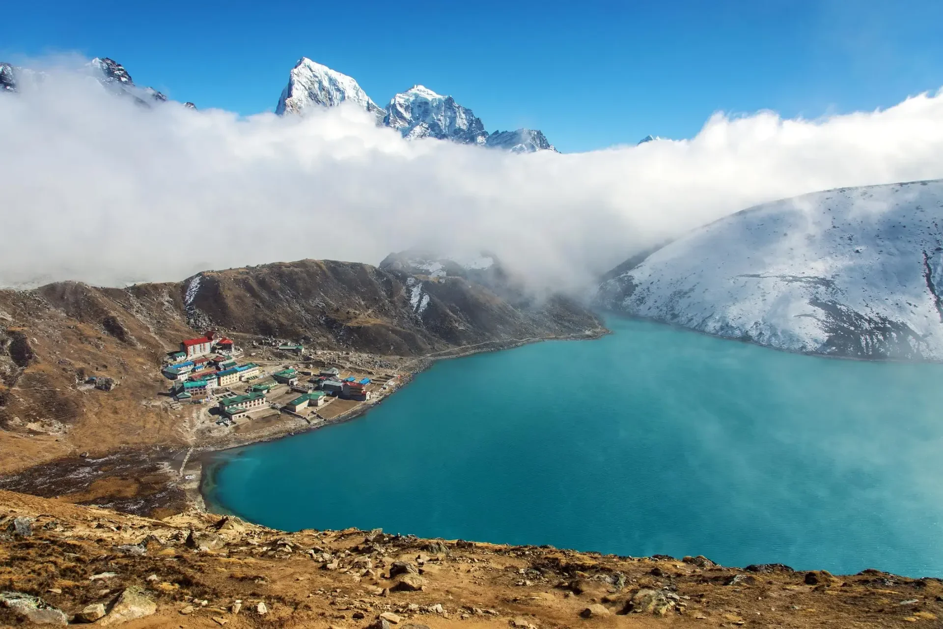 High-angle panoramic view of the turquoise Gokyo Lake and village partially covered by moving low clouds with snowy Himalayan peaks in the background