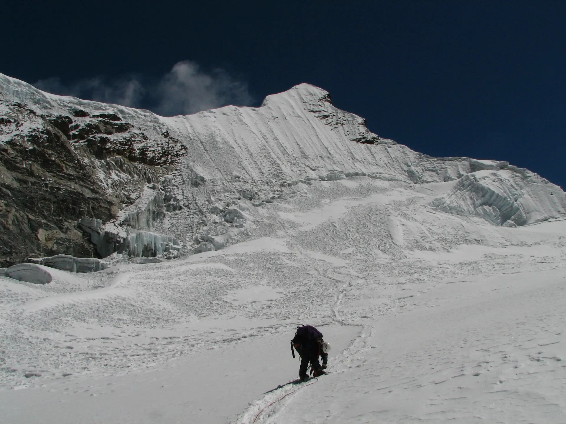 Mountaineer climbing steep snow slope towards rugged mountain peak under clear blue sky