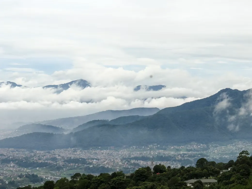 An aerial, high-angle view looking down at the sprawling Kathmandu city valley nestled below lush green hills and a thick layer of white clouds.