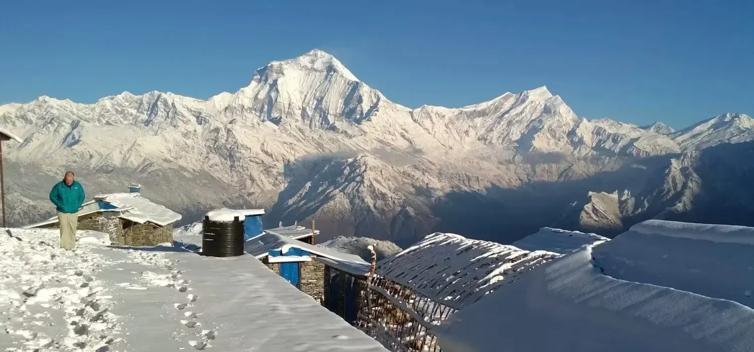 A trekker standing near snow-covered stone lodges at Khopra Danda showing the majestic, snow-capped Mount Dhaulagiri in the Annapurna region of Nepal.