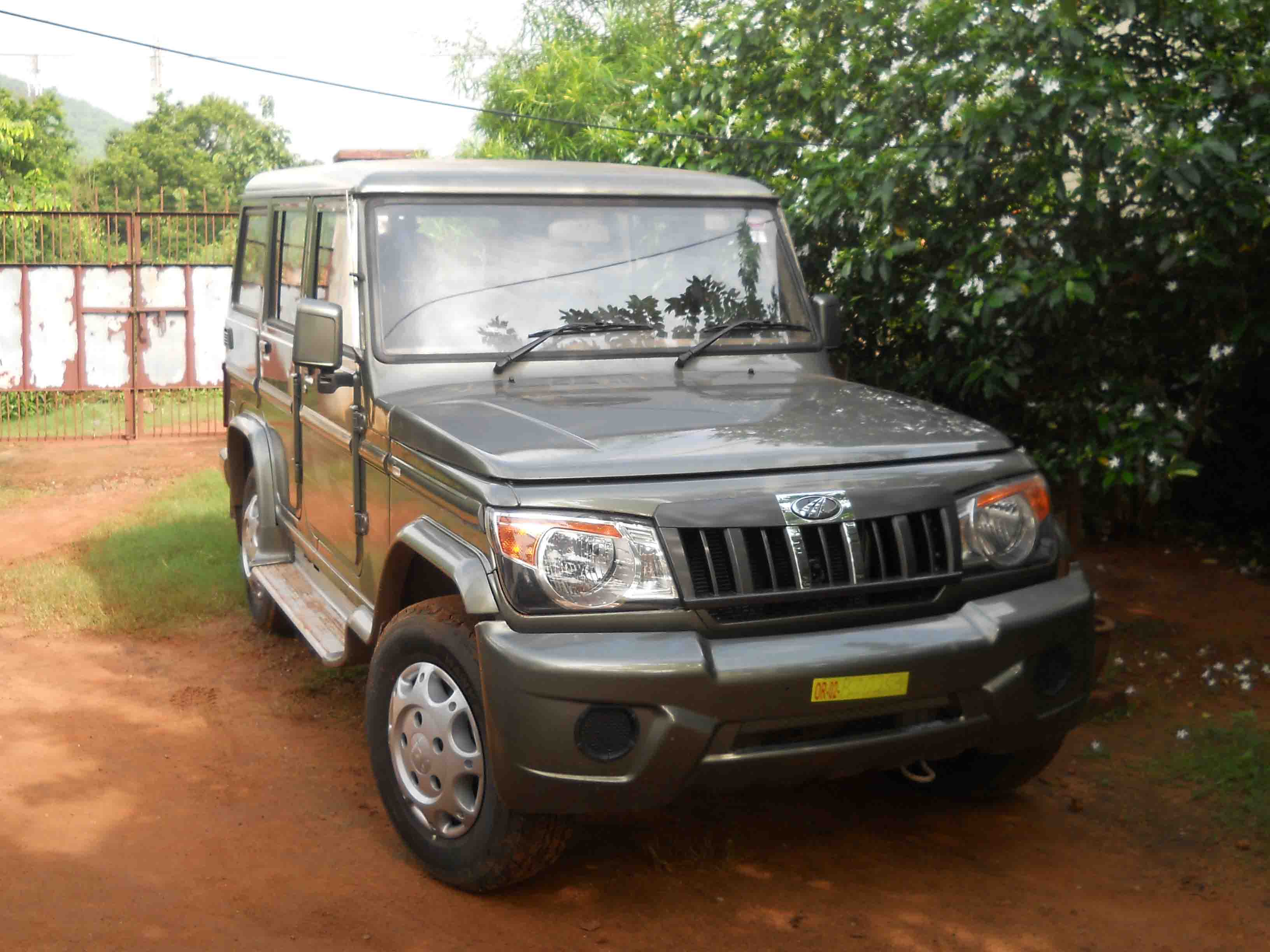 Front three-quarter view of a grey Mahindra Bolero rugged utility vehicle parked on a dirt surface next to green trees and an iron gate.