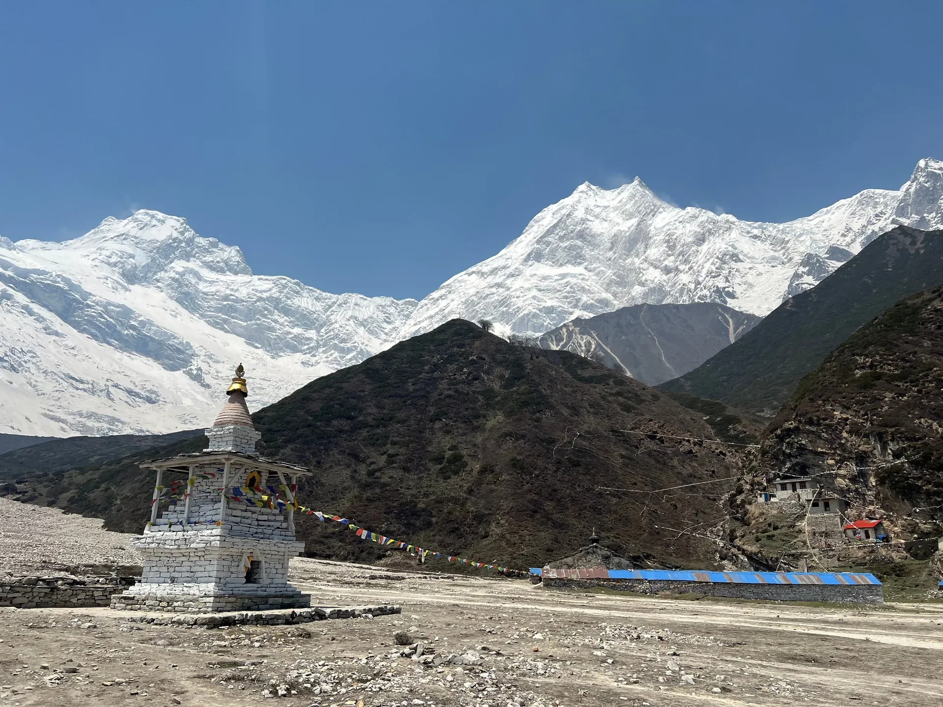A traditional white stone Buddhist chorten adorned with prayer flags in front of towering snow-capped peaks on the Manaslu Circuit trek
