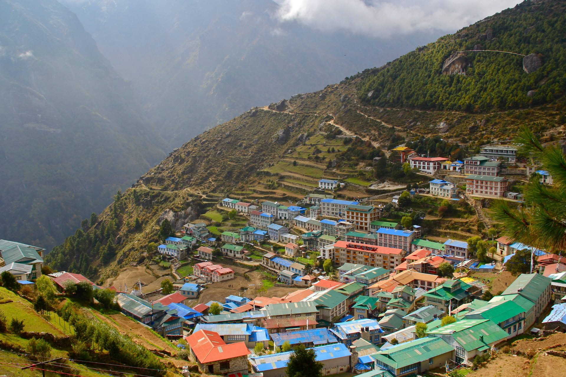 A colorful horseshoe-shaped village built into the steep green terraced hillsides of Namche Bazaar, surrounded by deep mountain valleys and hazy, mist-covered ridges in the Khumbu region.