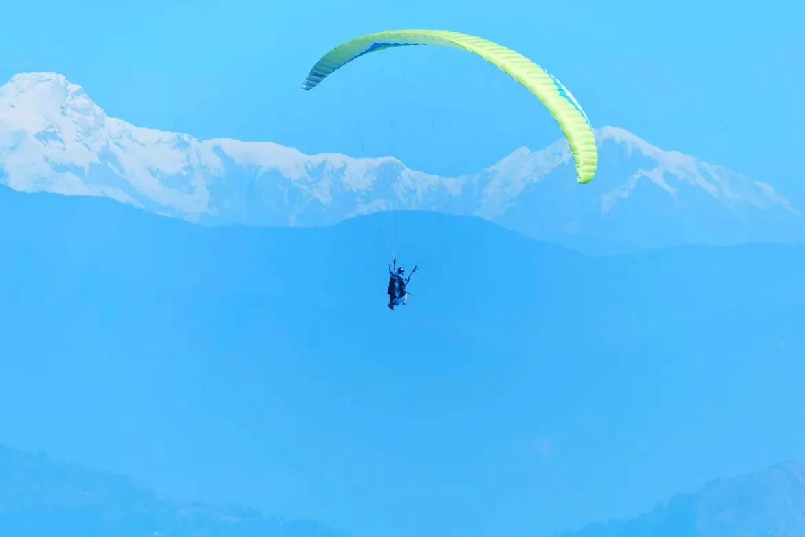 Tandem paraglider soaring through the clear blue sky with a bright yellow-green canopy directly in front of the snow-capped Annapurna mountain range in Pokhara.