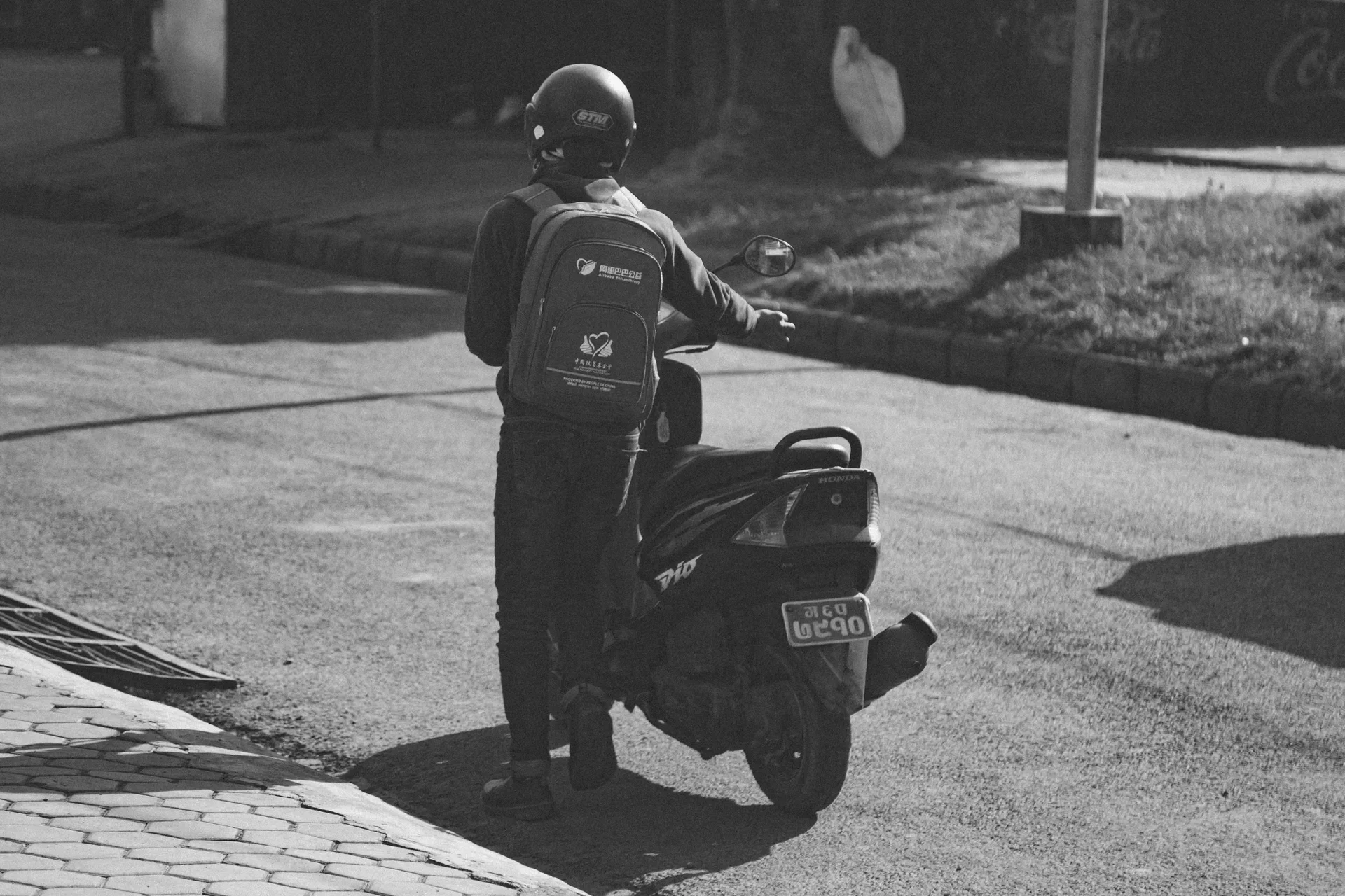 Black and white shot of a person wearing a helmet and backpack standing next to a Honda Dio scooter on a street in Pokhara