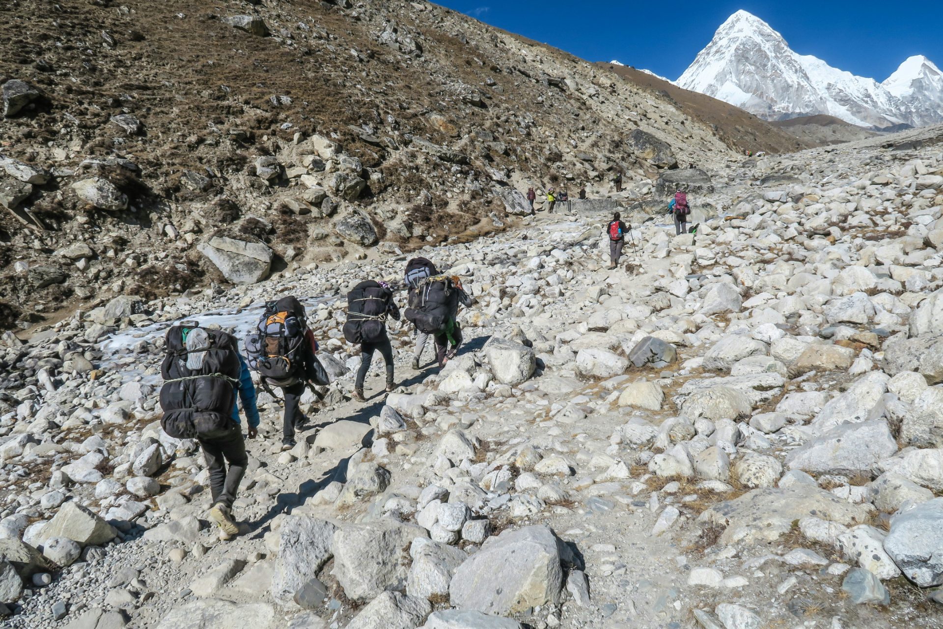 A line of local porters and Sherpas carrying heavy backpacks trekking up a rocky, boulder-strewn trail in the Khumbu region of Nepal toward a distant snow mountain.