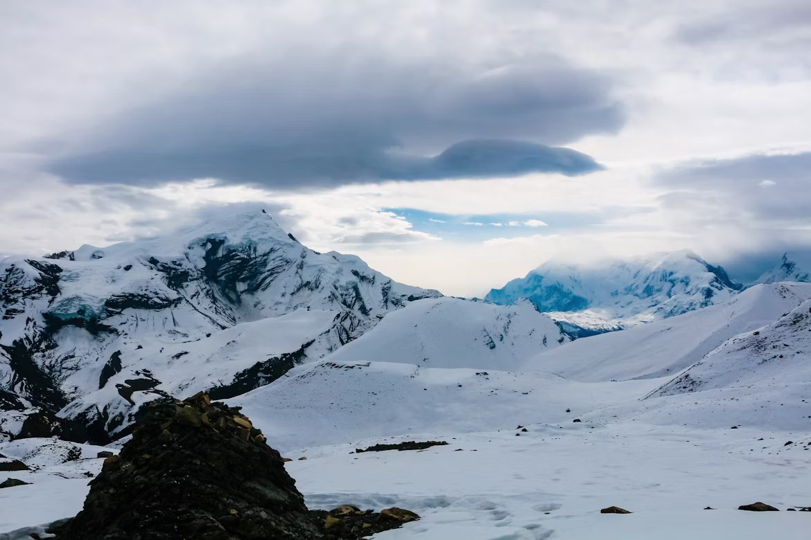 A snow-covered high-altitude mountain landscape under a heavy, dramatic storm cloud during a trek near Thorong La Pass.
