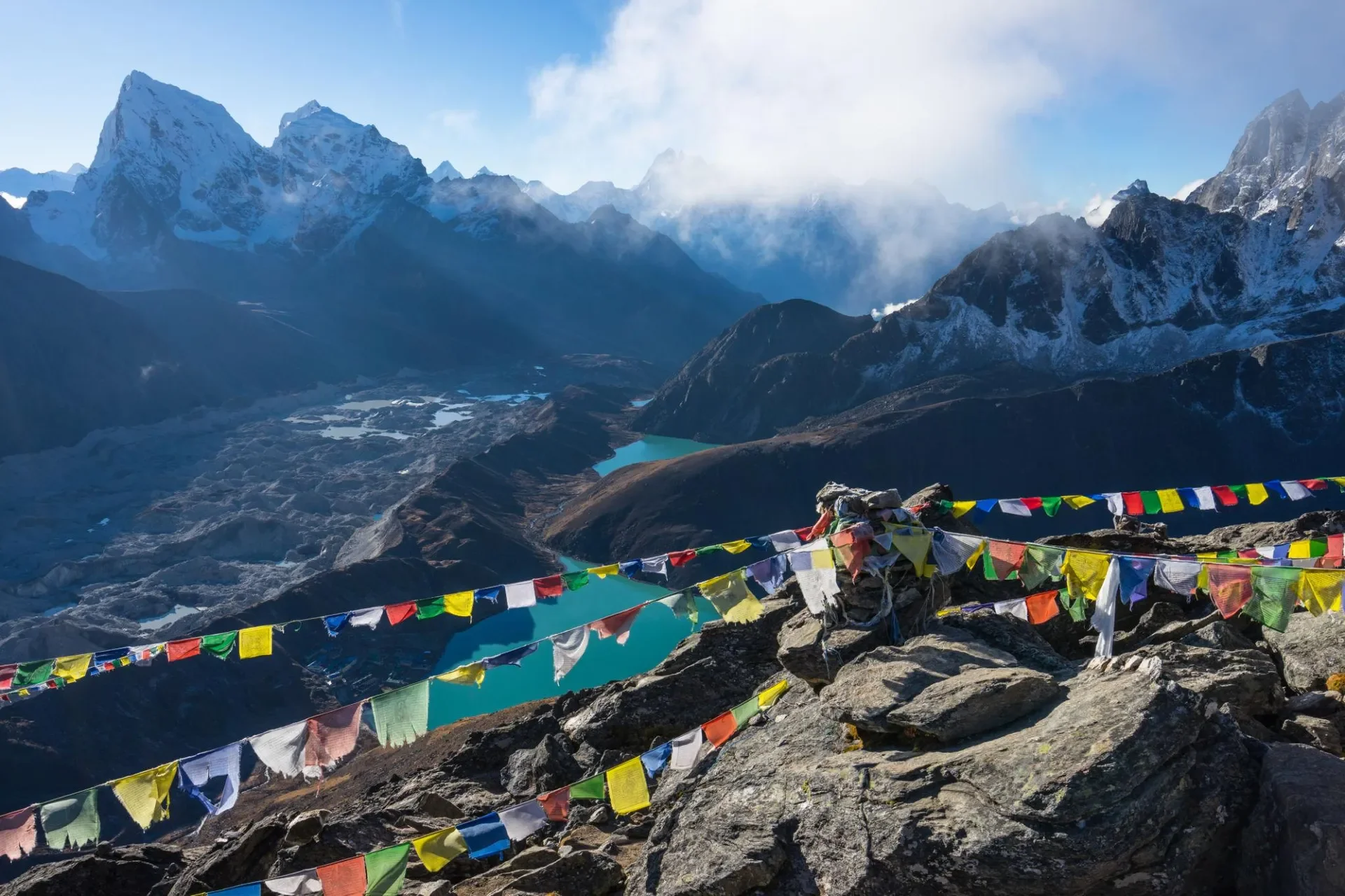 Looking down from the summit of Gokyo Ri decorated with colorful Buddhist prayer flags, revealing the vast Ngozumba Glacier and turquoise lakes below