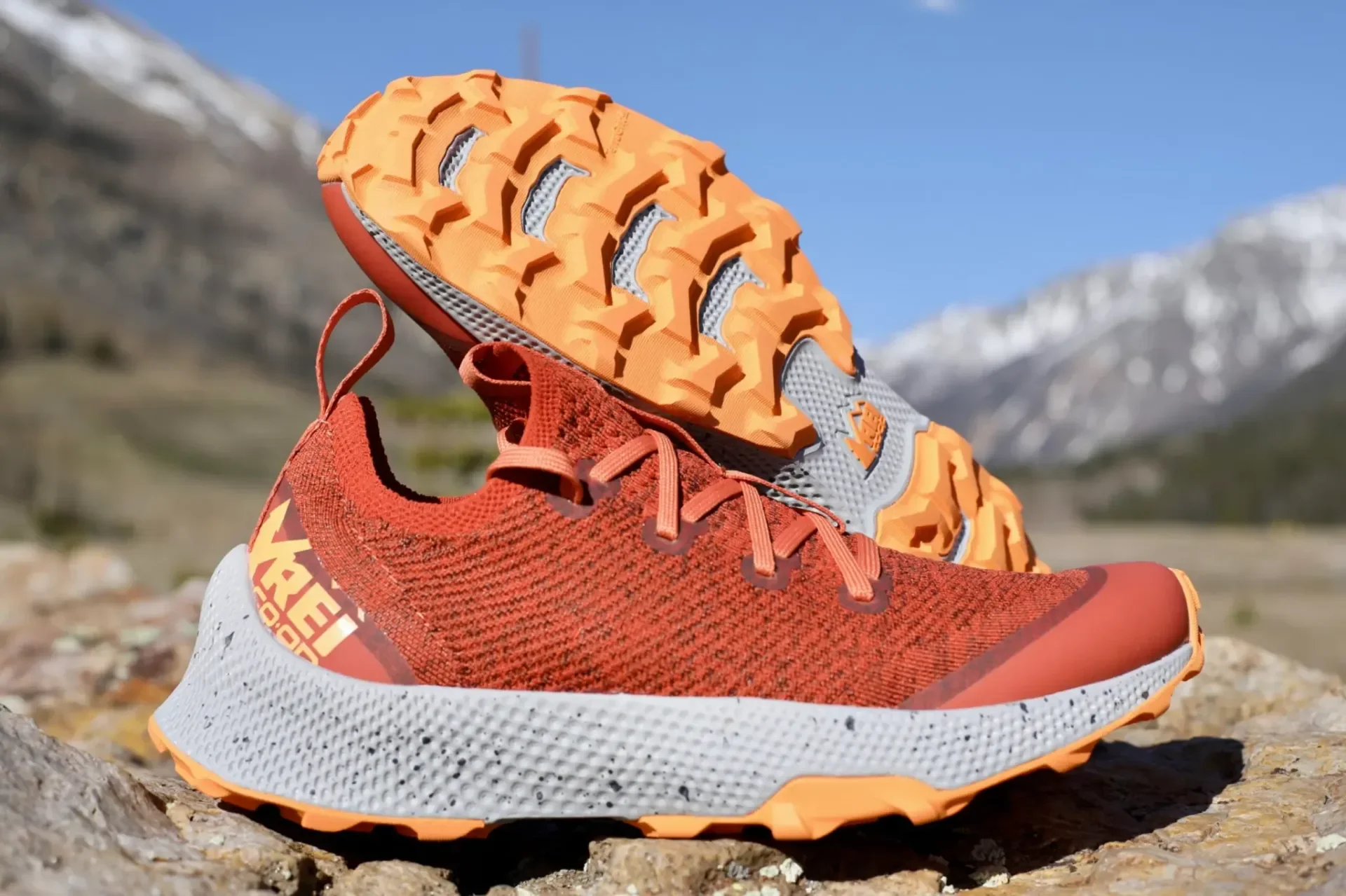 An orange REI Co-op trail running shoe displayed on a rock showcasing its aggressive, deep-lugged outsole with mountains in the background