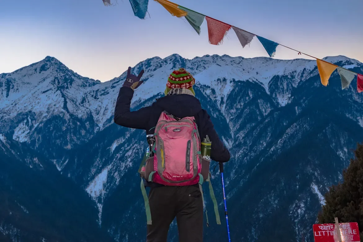 A hiker wearing a pink backpack gesturing with hand signs towards snow-capped mountain ridges under colorful Buddhist prayer flags on the Gosaikunda trekking route.