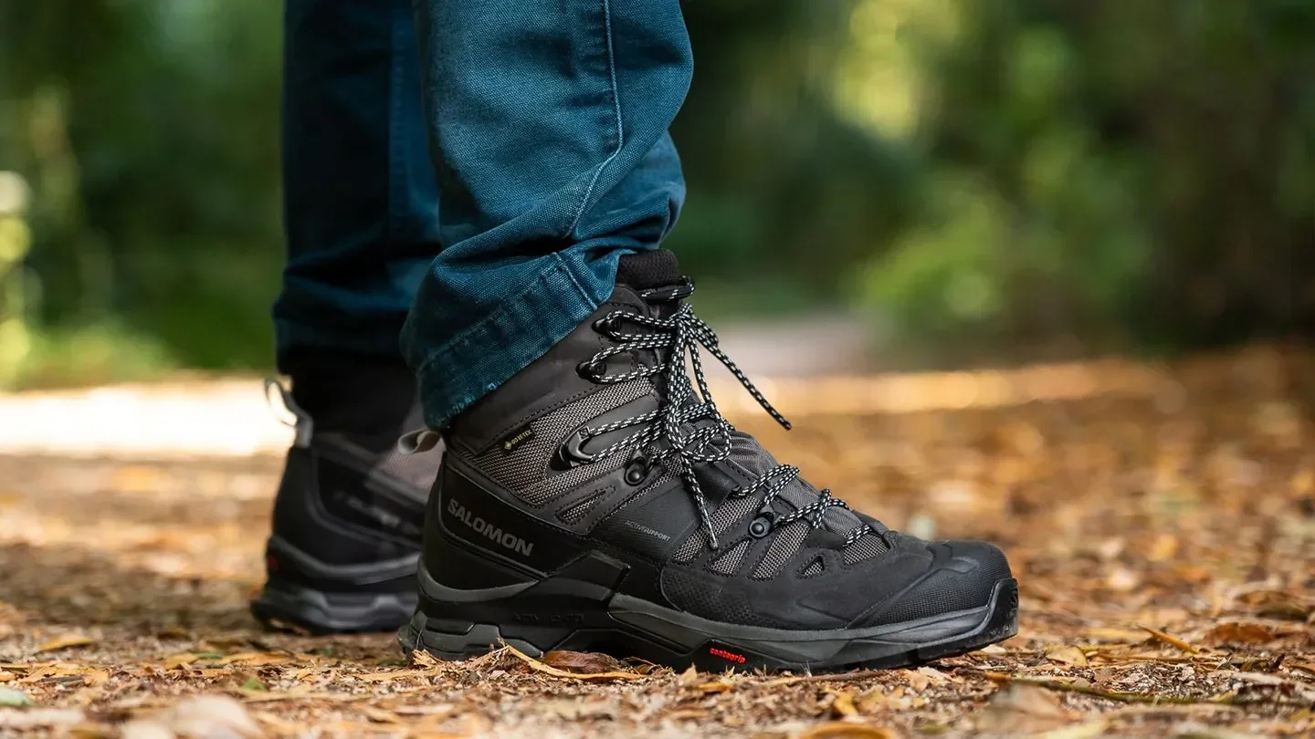 Side view of a hiker wearing dark grey Salomon mid-cut trekking boots and blue jeans on a leaf-covered forest trail