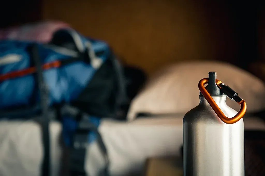 Close-up of a silver aluminum reusable water bottle with an orange carabiner clip on a bedside table with a blurred blue trekking backpack in the background