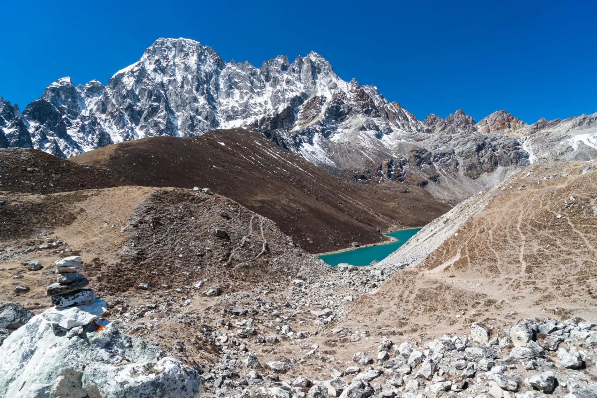 A rocky trekking path marked with a stone cairn leading toward a turquoise lake tucked between rugged, snow-dusted mountain ridges